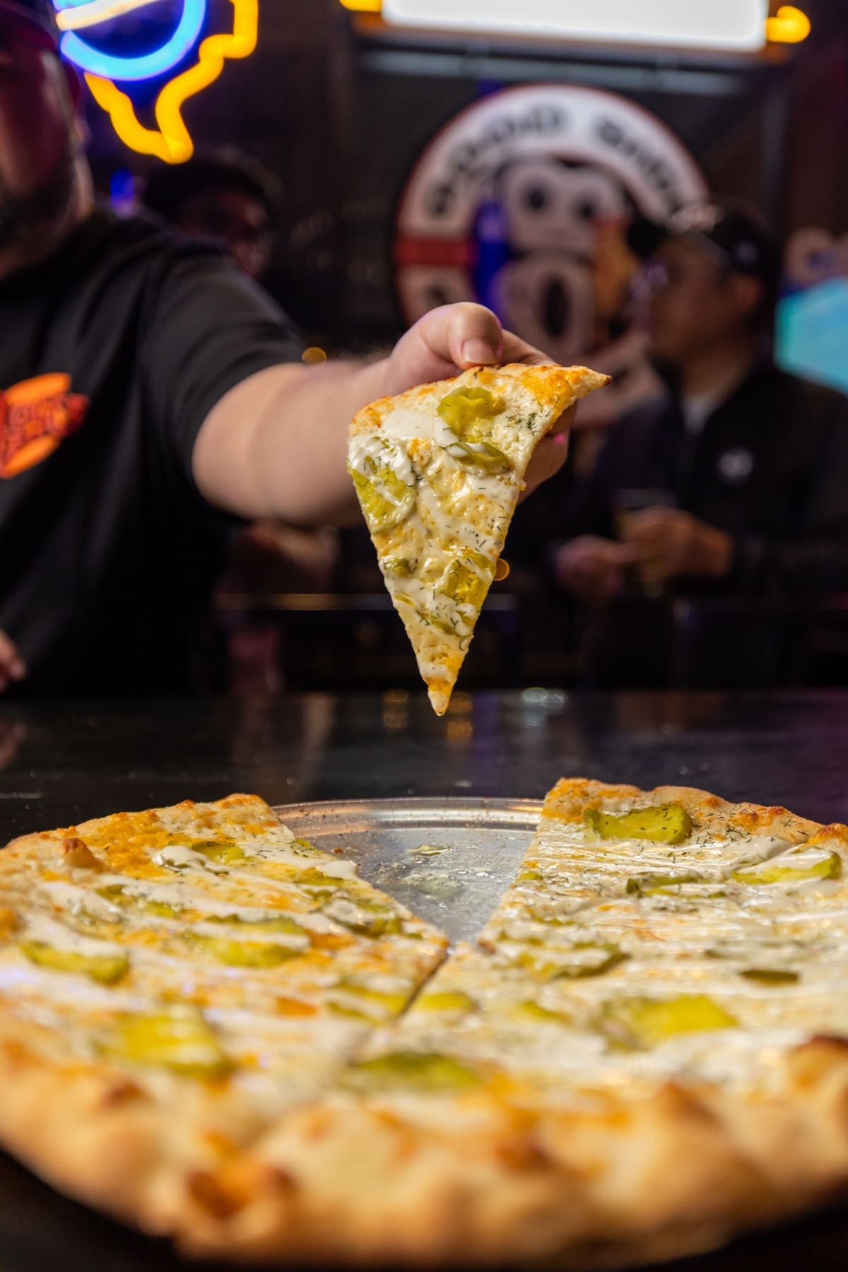 A slice of pickle pizza held up over the pie, the bar behind it